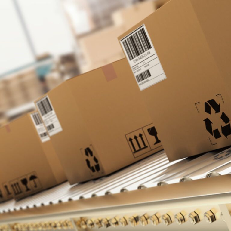 Several cardboard boxes with barcodes and shipping symbols move along a conveyor belt in a warehouse or distribution center, suggesting a shipping or logistics process.