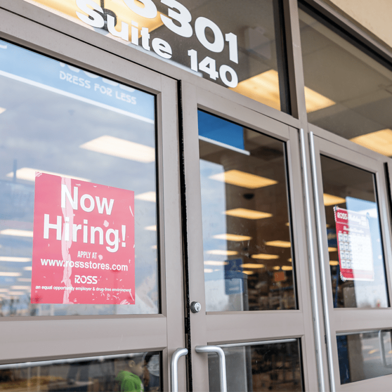 A glass storefront door displays a red “Now Hiring!” sign with application details for Ross. The store’s interior is visible through the windows, showing fluorescent lights and merchandise.