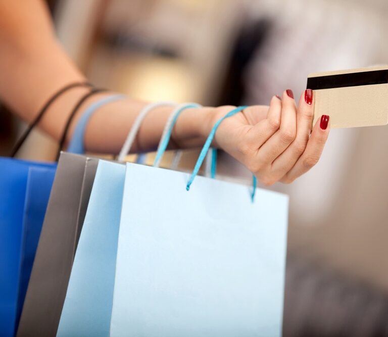 A person with red-painted nails holds several shopping bags and a credit card, suggesting a shopping activity. The background is blurred, keeping the focus on the hand, bags, and card.