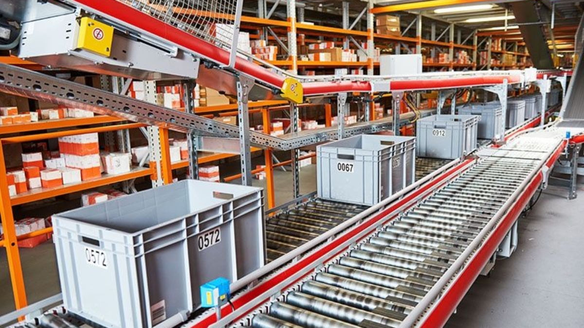 Plastic bins labeled with numbers move along a conveyor belt in a warehouse filled with shelves of orange and white packages. The setting appears organized and industrial, suggesting a logistics or distribution center.