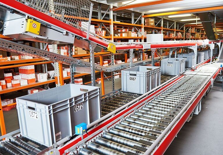 Plastic bins labeled with numbers move along a conveyor belt in a warehouse filled with shelves of orange and white packages. The setting appears organized and industrial, suggesting a logistics or distribution center.