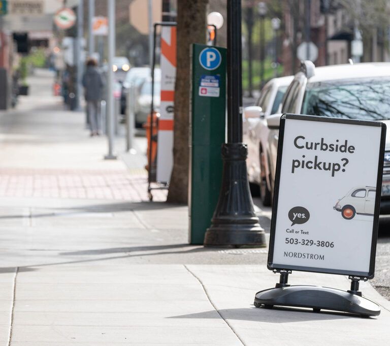 A sidewalk sign reads “Curbside pickup? yes” with a phone number and a small car graphic, promoting Nordstrom’s curbside service. Cars are parked on the street and pedestrians walk in the background.