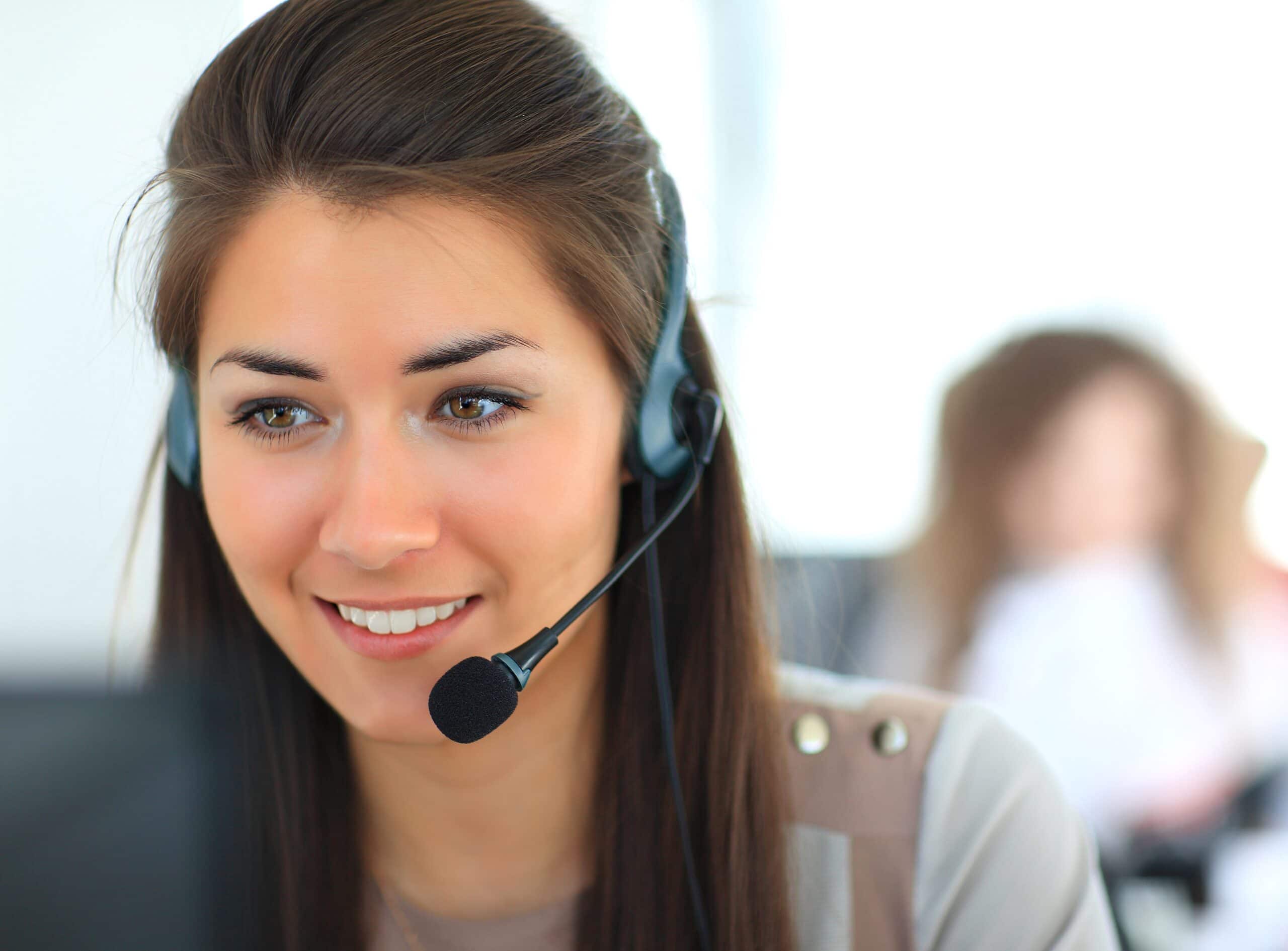 A woman with long brown hair wearing a headset smiles while working at a computer in an office. Another person is blurred in the background.