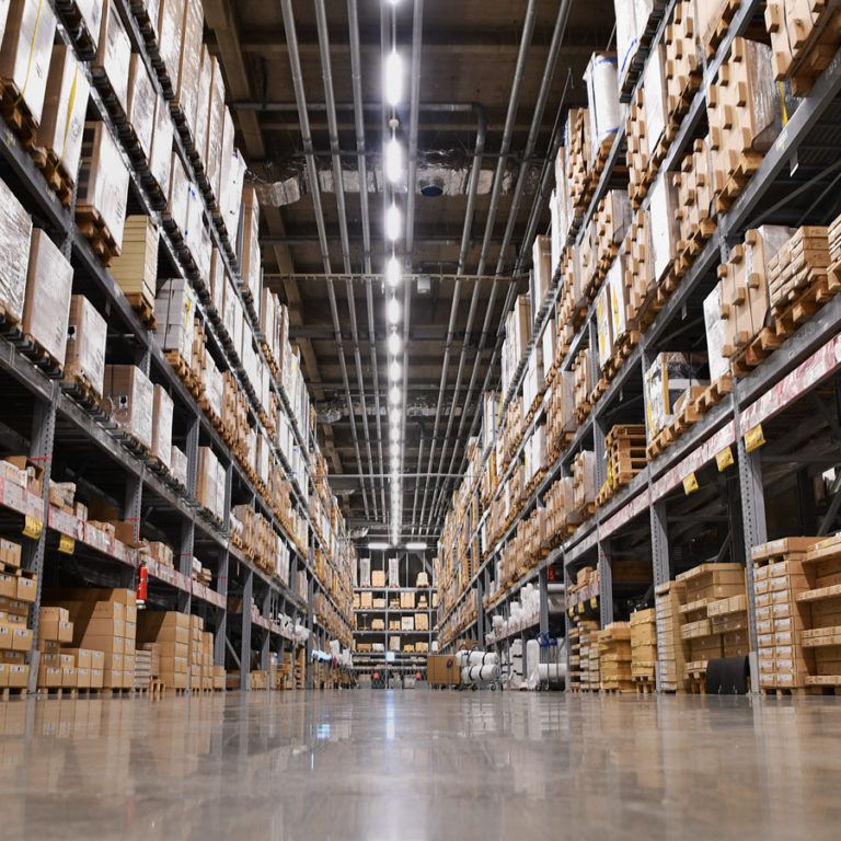 View of a large warehouse with tall shelves stacked with cardboard boxes, ideal for warehouse capacity analysis, bright overhead lighting, and a shiny concrete floor, seen from a low angle between the aisles.