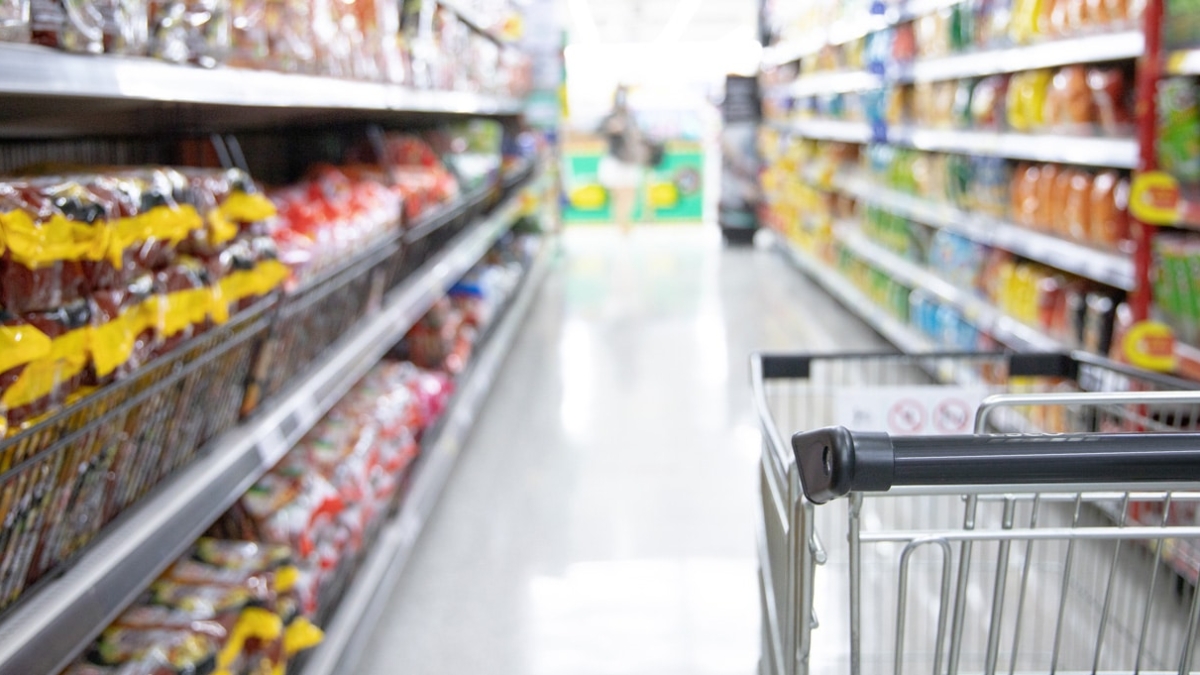 A shopping cart in a grocery store aisle, surrounded by shelves stocked with various colorful packaged food items. The image is bright and the shelves are well-organized.
