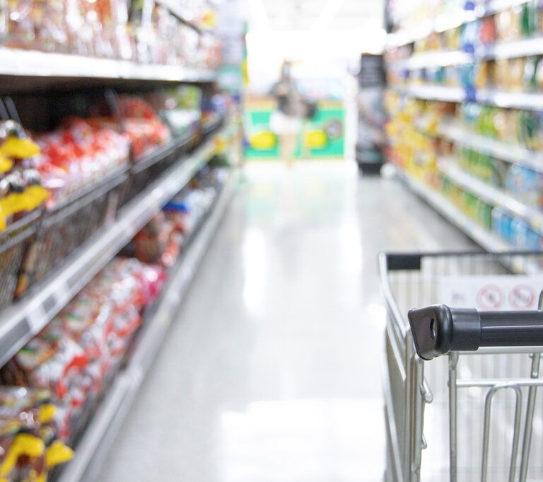 A shopping cart in a grocery store aisle, surrounded by shelves stocked with various colorful packaged food items. The image is bright and the shelves are well-organized.