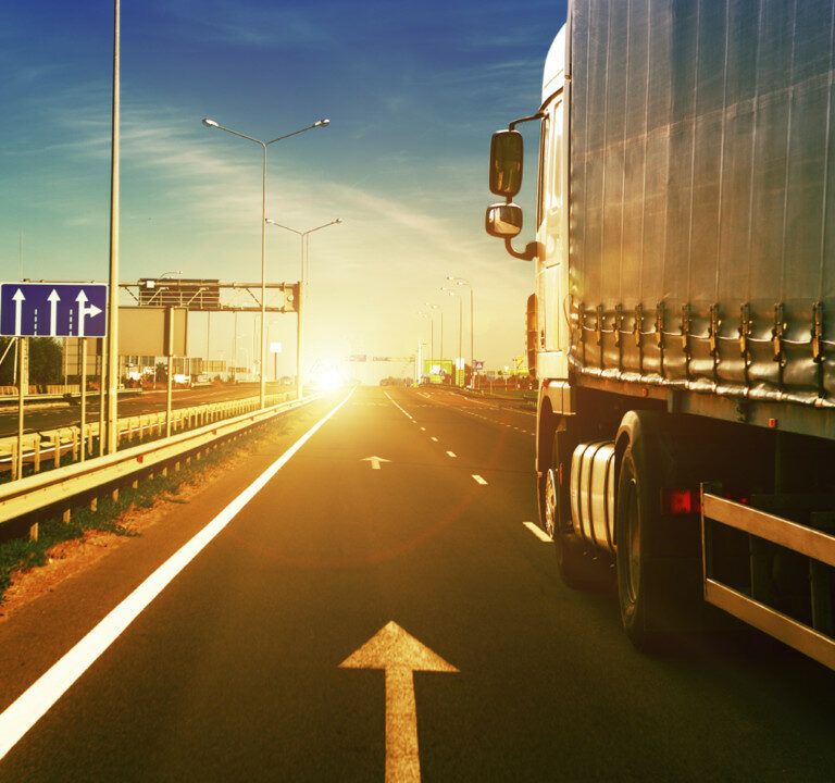 A large truck drives along a highway at sunset, with sunlight illuminating the road ahead. The highway has clear lane markings, road signs, guardrails, and a few vehicles in the distance.