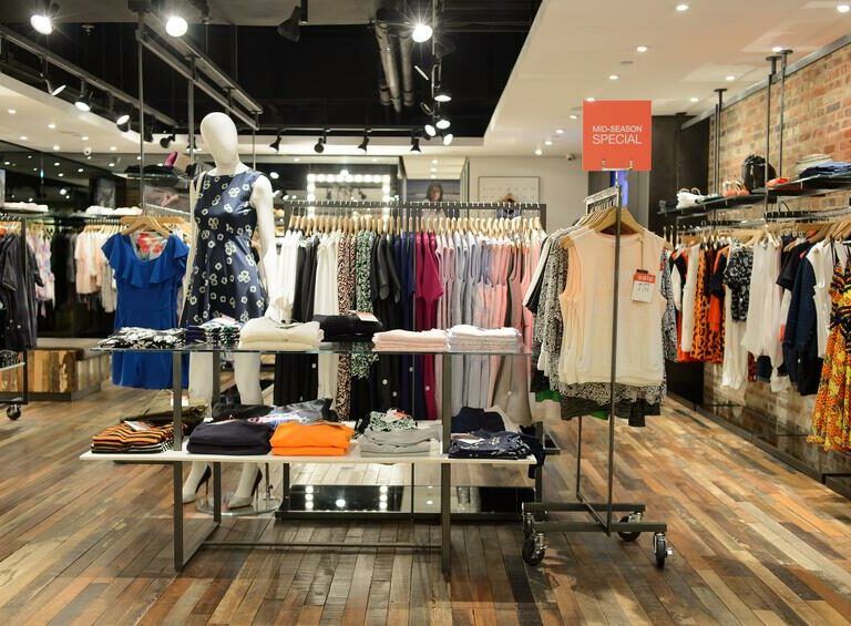 Clothing store interior with wooden floors, racks of colorful dresses and shirts, a mannequin in a blue dress, folded clothes on tables, and a sign that reads Mid-Season Special. Lean retail principles shine in the bright, modern lighting overhead.