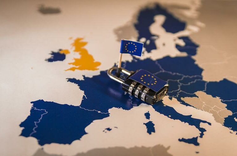 A padlock with a small European Union flag is placed on a map of Europe, highlighting EU member countries in blue and the UK in yellow, symbolizing security or Brexit.
