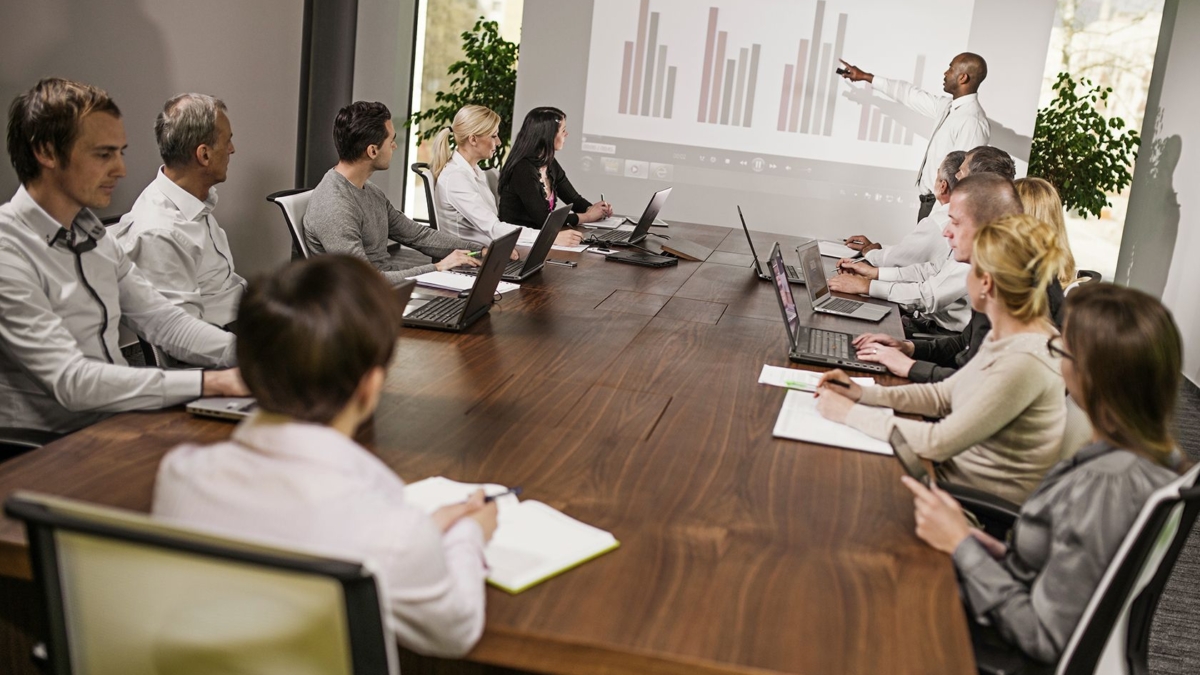 A group of people in business attire sit around a conference table with laptops and notepads, watching a presenter point to a bar chart on a projected screen during a meeting about the Microsoft Dynamics AX upgrade.