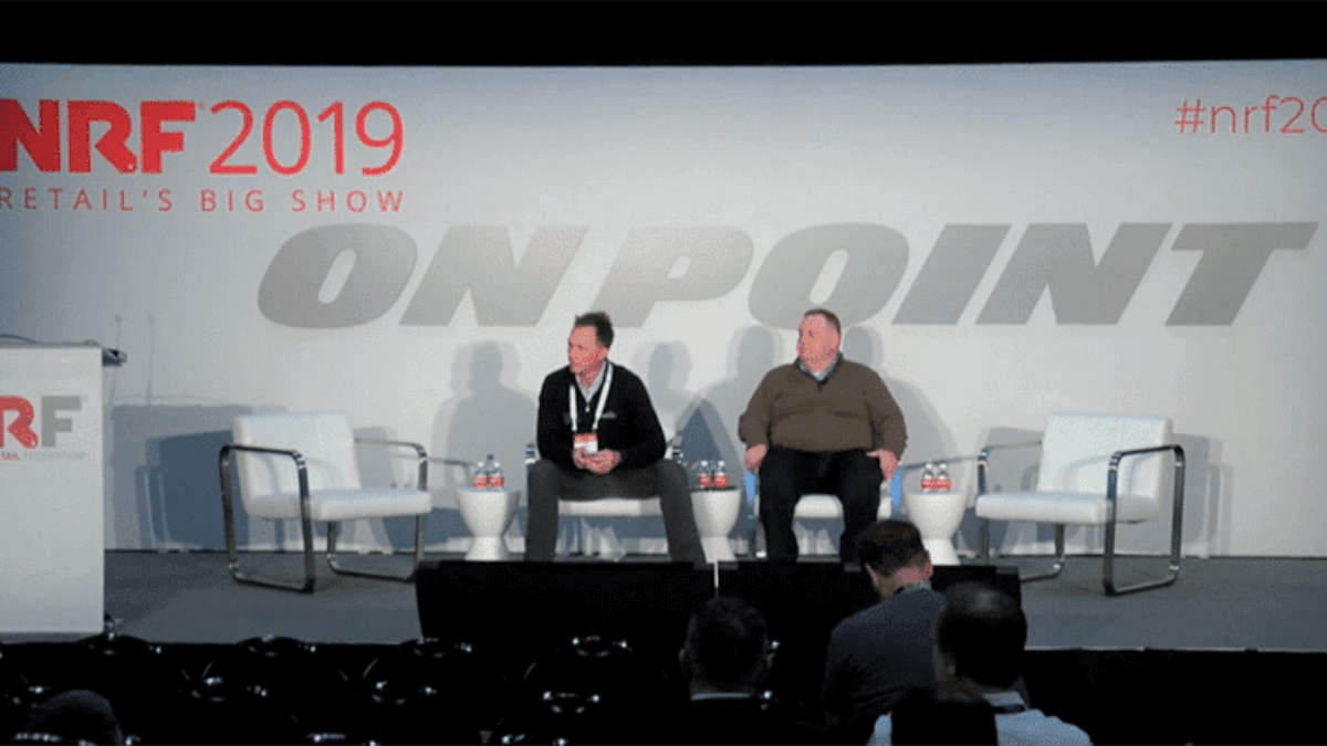 Two men sit on a stage with three empty chairs at NRF 2019 Retail’s Big Show. The backdrop has “ON POINT” and event branding. A podium is on the left, and a few people are seated in the audience.