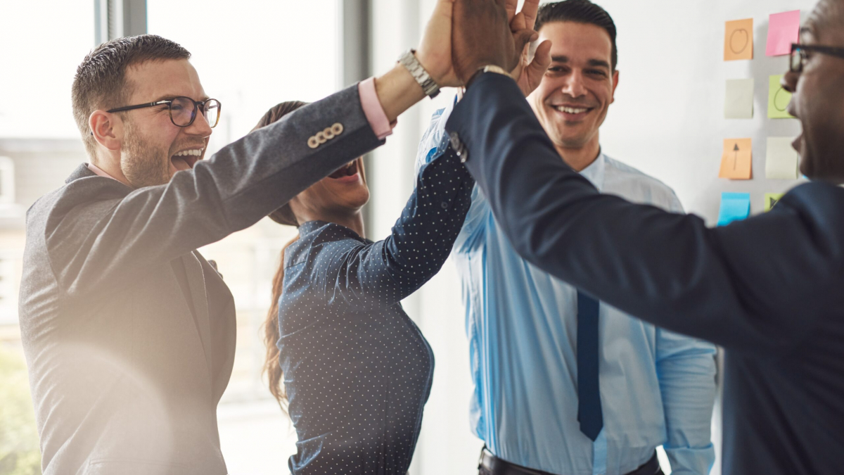 Four business professionals, three men and one woman, smiling and giving a group high-five in an office setting with sticky notes on the wall in the background.