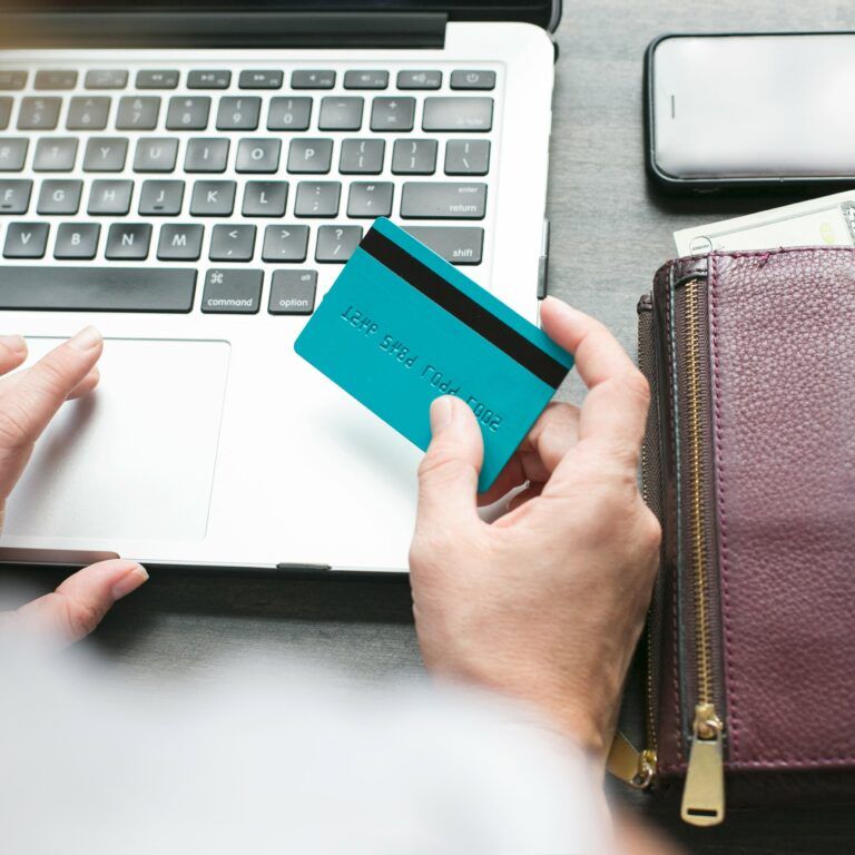 A person uses a laptop while holding a blue credit card; a purple wallet with cash, coins, and a smartphone are nearby on the table.