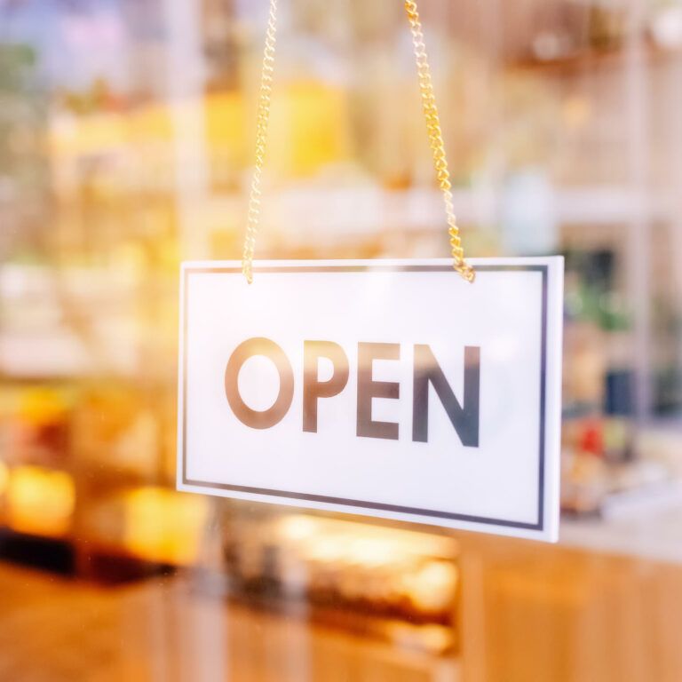 A white sign with the word OPEN in bold black letters hangs on a glass door, with a blurred view of a brightly lit interior in the background.