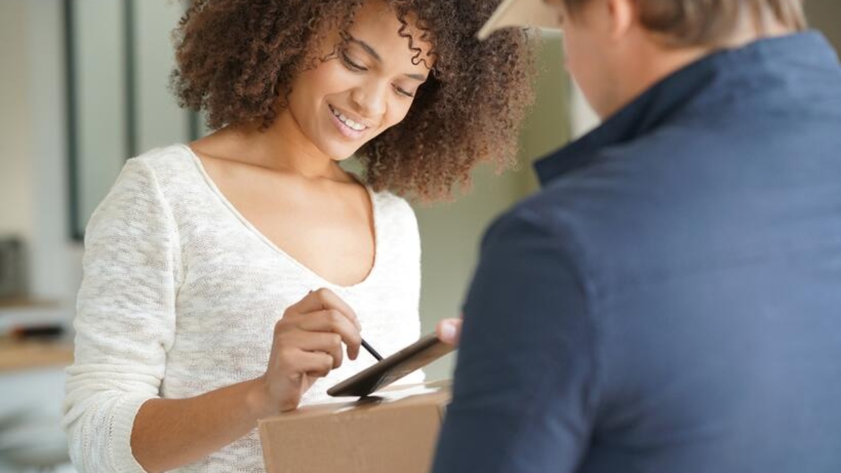 A woman smiles as she receives a package from a delivery person and signs for it on a digital device.