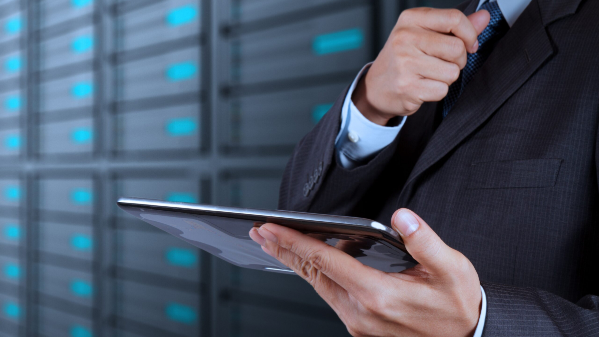 A person in a suit adjusts their tie while holding a tablet, standing in front of a blurred background of server racks in a data center.