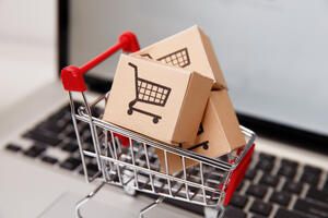 A miniature shopping cart holds small cardboard boxes with shopping cart icons, placed on a laptop keyboard, symbolizing online shopping or e-commerce.
