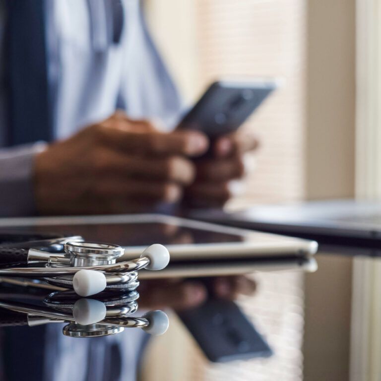A stethoscope rests on a desk near a tablet and laptop, while a person in business attire uses a smartphone in the background, suggesting a medical or healthcare professional at work.
