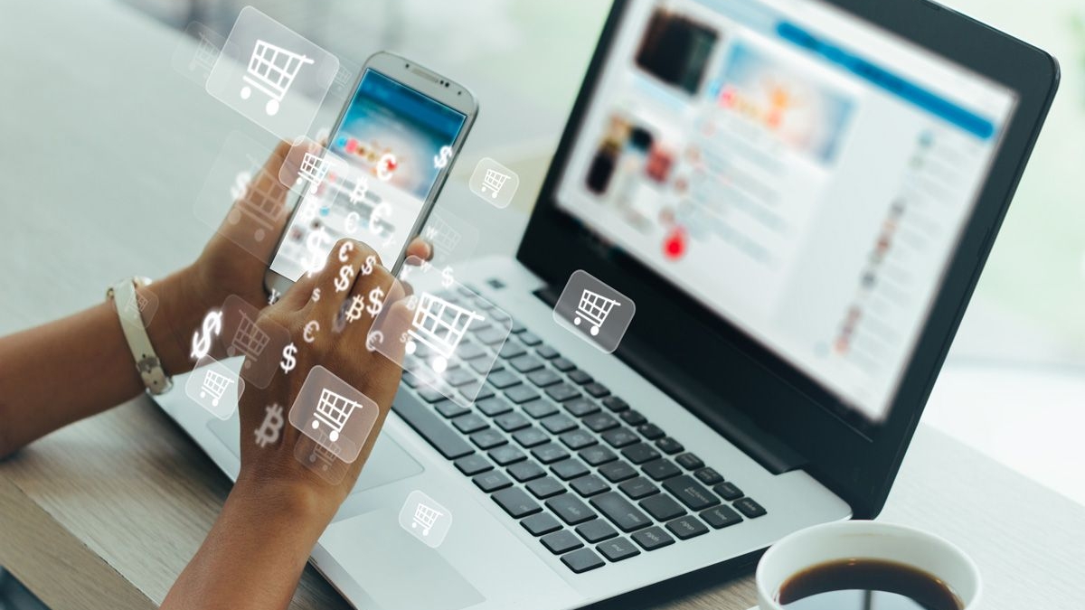 A person uses a smartphone and laptop at a table with a cup of coffee. Digital shopping cart and currency icons are overlaid, suggesting online shopping or e-commerce activity.