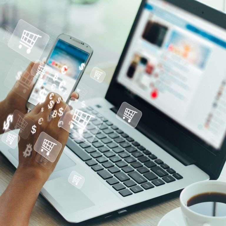 A person uses a smartphone and laptop at a table with a cup of coffee. Digital shopping cart and currency icons are overlaid, suggesting online shopping or e-commerce activity.