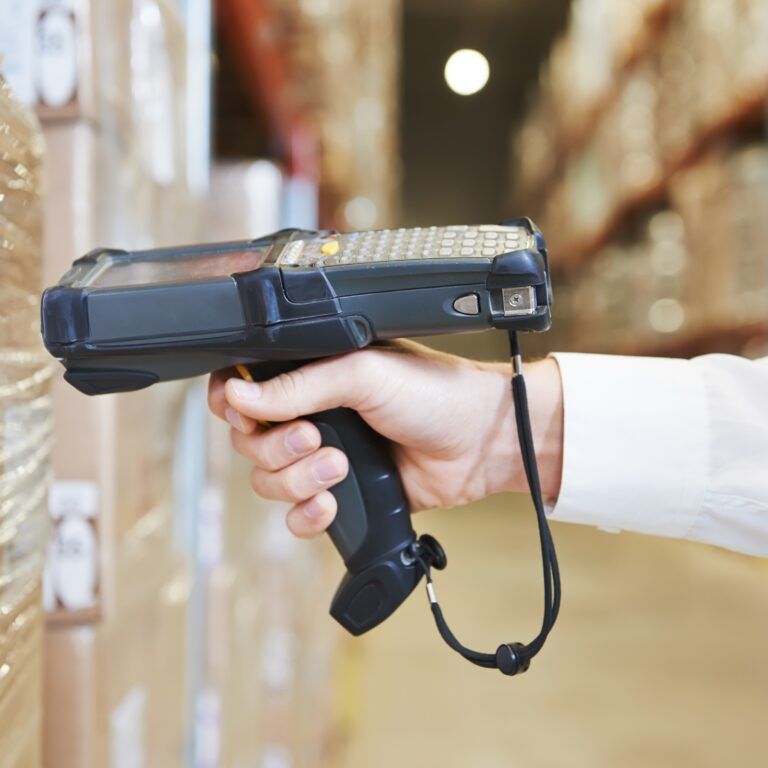 A person scans a barcode on a pallet of boxes in a warehouse using a handheld barcode scanner, helping ensure food traceability. Shelves filled with products are visible in the background.