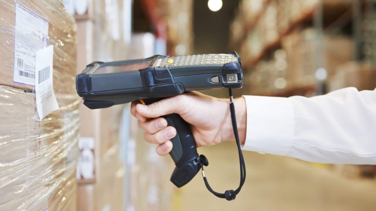 A person scans a barcode on a pallet of boxes in a warehouse using a handheld barcode scanner, helping ensure food traceability. Shelves filled with products are visible in the background.