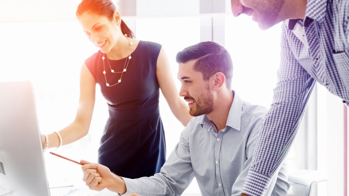 Three colleagues smile and collaborate at a desk, looking at a computer screen. One woman stands pointing at the monitor, while two men sit and stand nearby, engaged in discussion in a bright office setting.