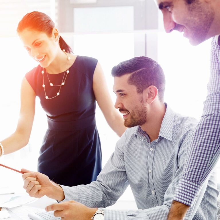 Three colleagues smile and collaborate at a desk, looking at a computer screen. One woman stands pointing at the monitor, while two men sit and stand nearby, engaged in discussion in a bright office setting.