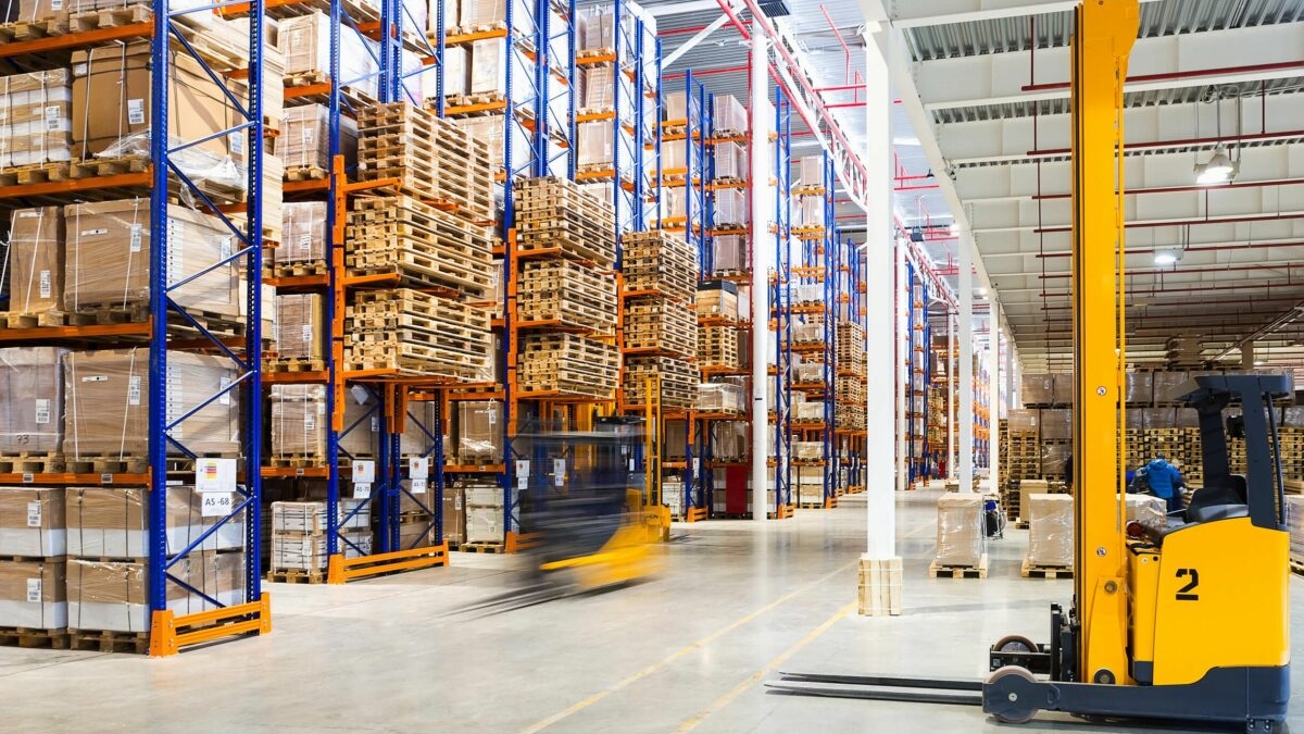 Warehouse with tall shelves stacked with boxes and pallets, a blurred forklift moving goods down the aisle, and another yellow forklift parked in the foreground; a bright and organized environment shows what slotting can achieve.
