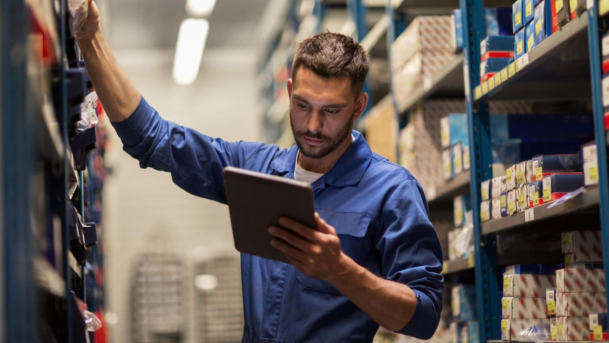 A man in a blue work jumpsuit stands between warehouse shelves, reaching for a box with one hand and holding a tablet in the other, checking inventory or stock.