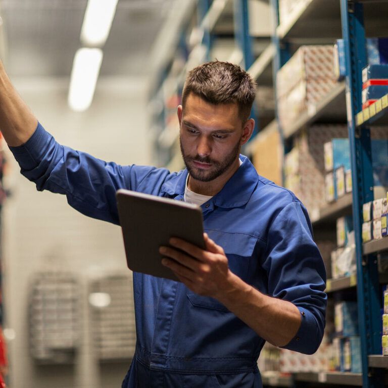A man in a blue work jumpsuit stands between warehouse shelves, reaching for a box with one hand and holding a tablet in the other, checking inventory or stock.