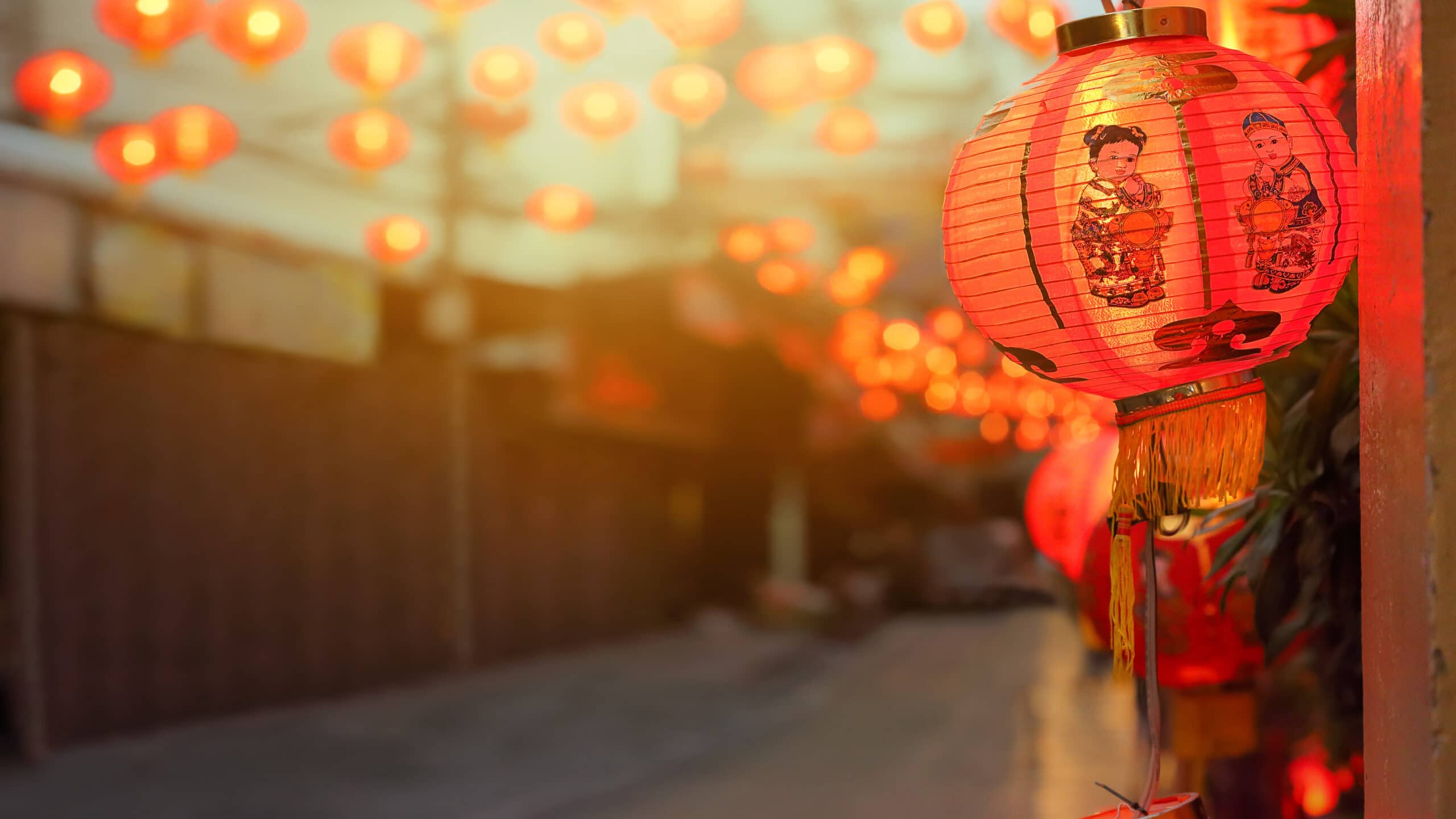 A red paper lantern with an illustration of a woman in traditional attire hangs outside, glowing among many similar lanterns lining a street in the evening. The background is softly blurred, emphasizing the lanterns’ warm light.