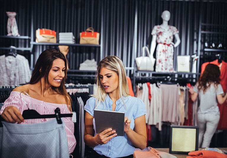 Two women are shopping for clothes in a store; one is holding a garment while the other shows her something on a tablet. Clothing and handbags are displayed in the background, with two more people browsing.