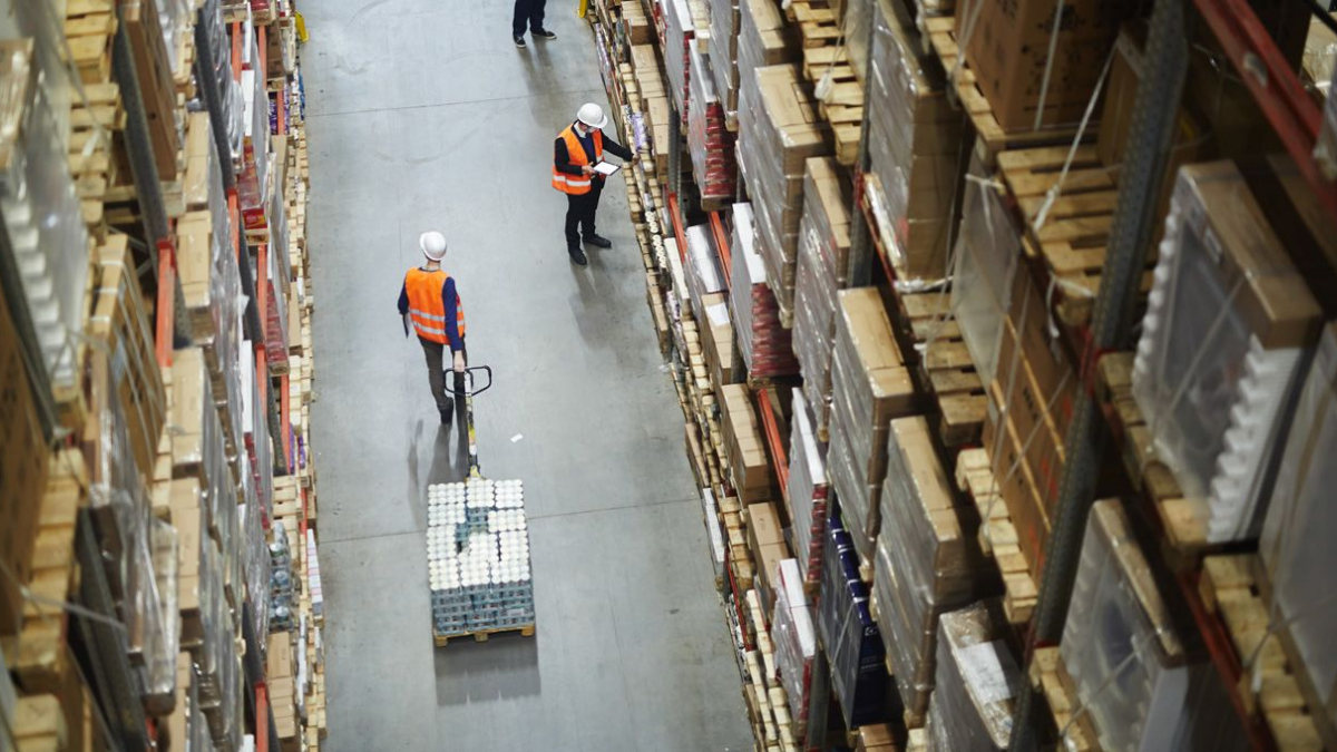 Workers in orange vests and helmets move among tall shelves stacked with boxes in a large warehouse. One worker uses a pallet jack to move goods, while another checks inventory with a clipboard.