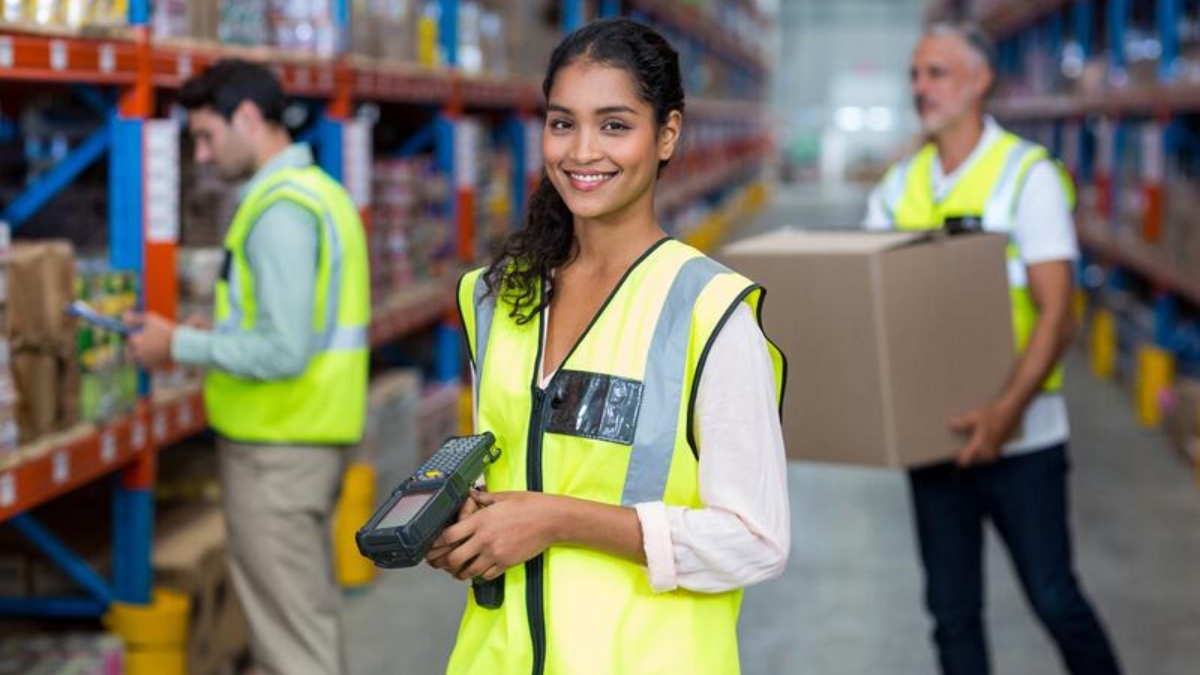 A woman in a yellow safety vest smiles at the camera while holding a barcode scanner in a warehouse. Two coworkers in safety vests work in the background, one handling a box and the other checking inventory.