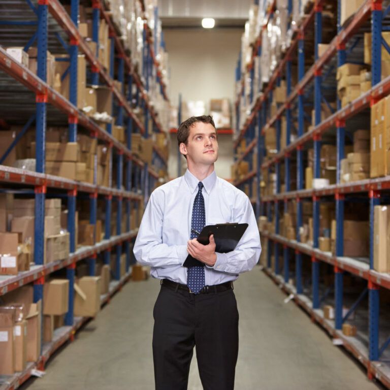 A man in business attire holding a clipboard stands in the aisle of a warehouse, surrounded by tall shelves filled with boxes and packages.