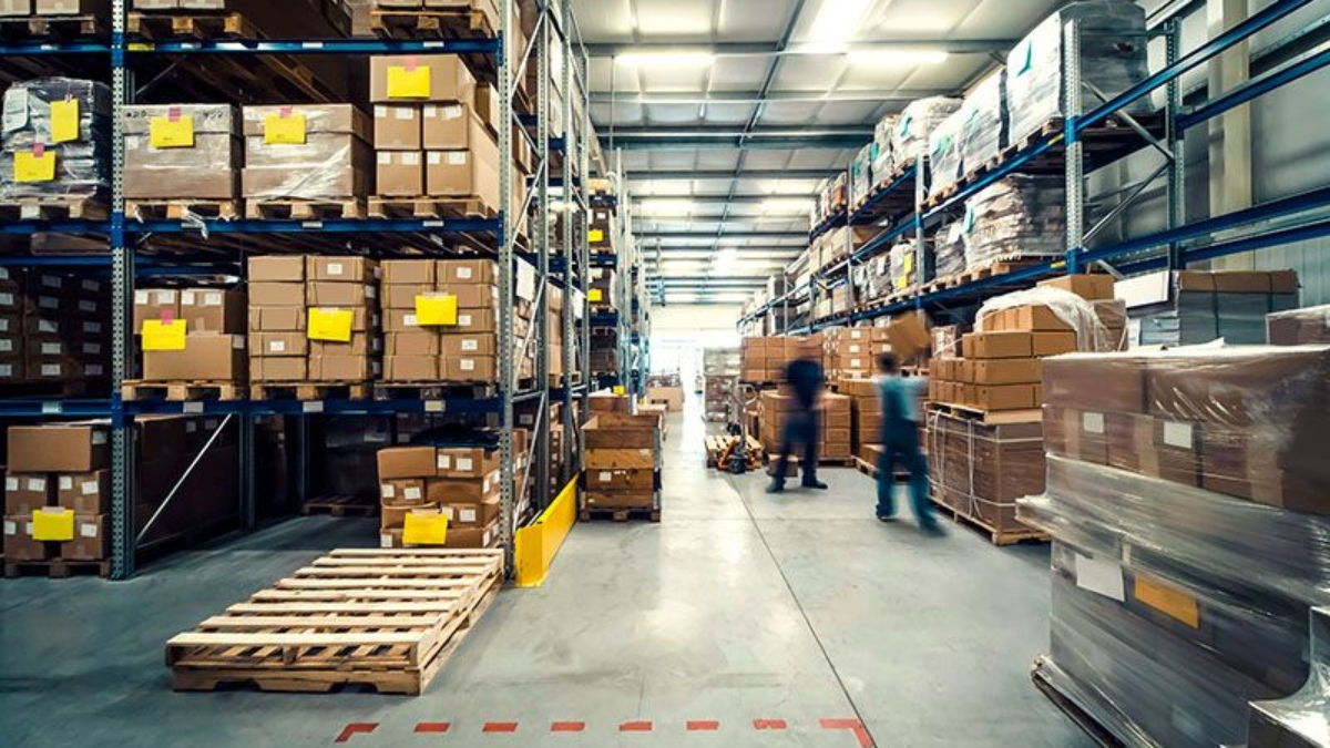 A spacious warehouse interior with tall racks filled with cardboard boxes. Two workers are moving among the shelves, and a wooden pallet is on the floor in the foreground. The scene is well-lit with overhead lights.