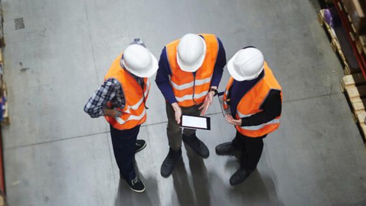 Three workers in orange safety vests and white hard hats stand in a warehouse, viewed from above, looking at a tablet together among shelves filled with boxes.