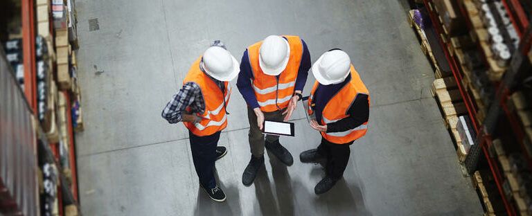 Three workers in orange safety vests and white hard hats stand in a warehouse, viewed from above, looking at a tablet together among shelves filled with boxes.