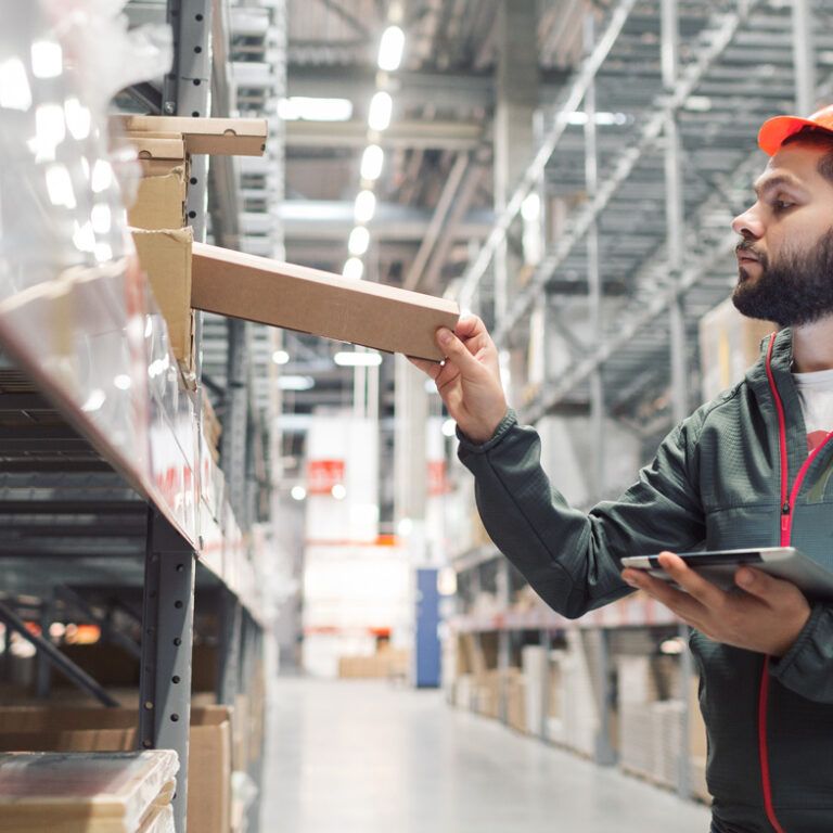 A man wearing an orange hard hat and dark jacket uses a tablet while placing a box on a shelf in a large warehouse with high racks and industrial lighting.