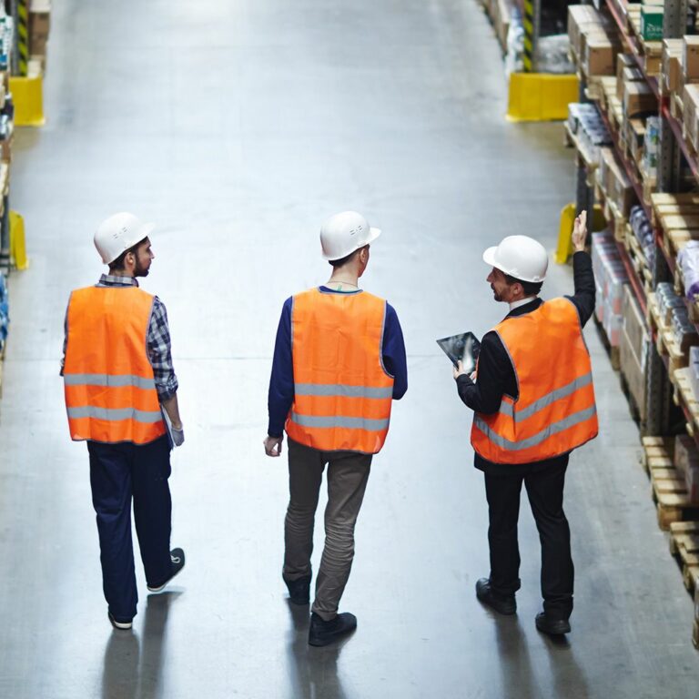 Three people wearing orange safety vests and white hard hats walk down an aisle in a large warehouse, surrounded by shelves stocked with various boxes and goods.