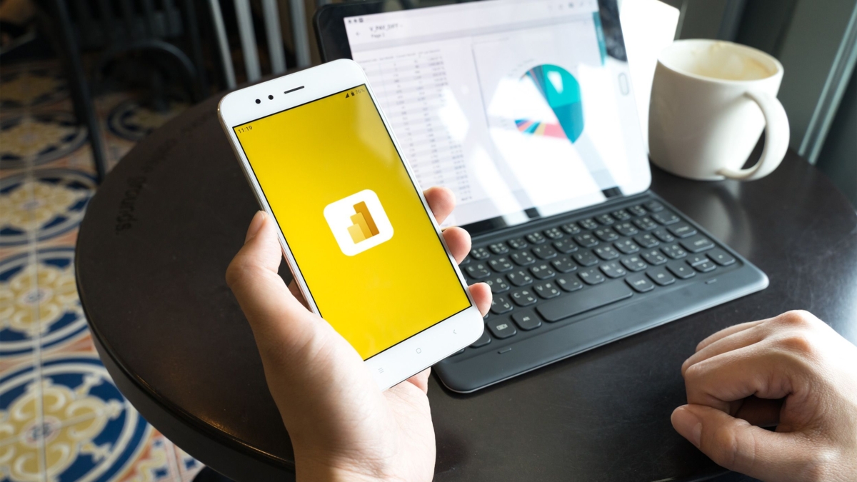 A person holds a smartphone displaying the Microsoft Power BI logo, while sitting at a round table with a tablet showing a colorful pie chart and a white coffee mug.