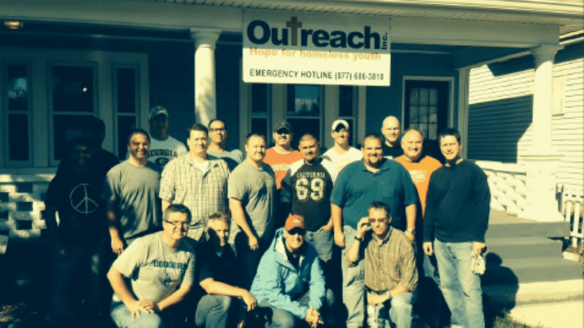 A group of fifteen men pose and smile together outside a house with a sign reading Outreach: Hope for Homeless Youth and an emergency hotline number above the porch.
