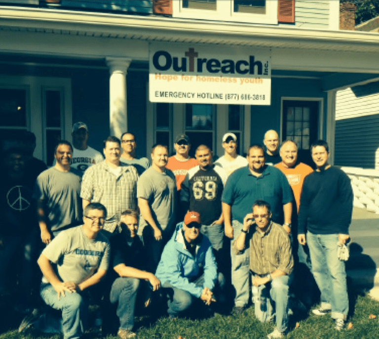 A group of fifteen men pose and smile together outside a house with a sign reading Outreach: Hope for Homeless Youth and an emergency hotline number above the porch.
