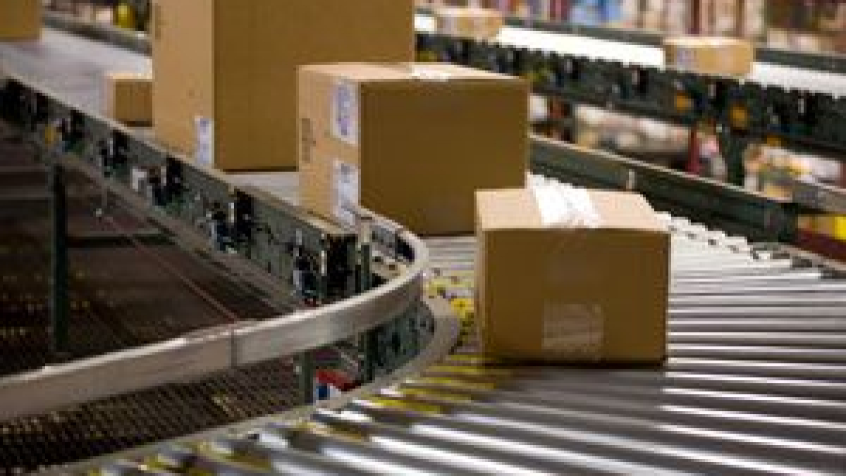 Several cardboard boxes travel along a curved conveyor belt in a warehouse or distribution center, surrounded by shelves stocked with other items.