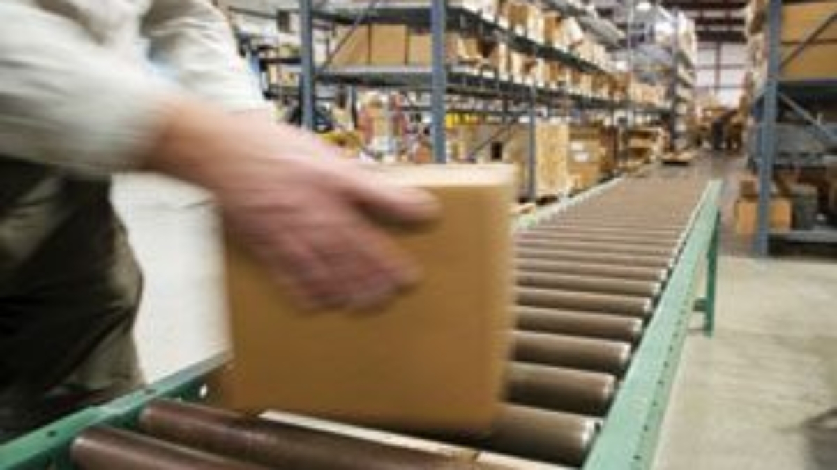 A person places a cardboard box on a conveyor belt in a warehouse with shelves full of boxes and packages in the background.