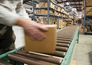 A person places a cardboard box on a conveyor belt in a warehouse with shelves full of boxes and packages in the background.