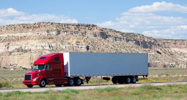 A red semi-truck with a white and gray trailer drives along a highway, with rocky, tan cliffs and a blue sky with scattered clouds in the background.