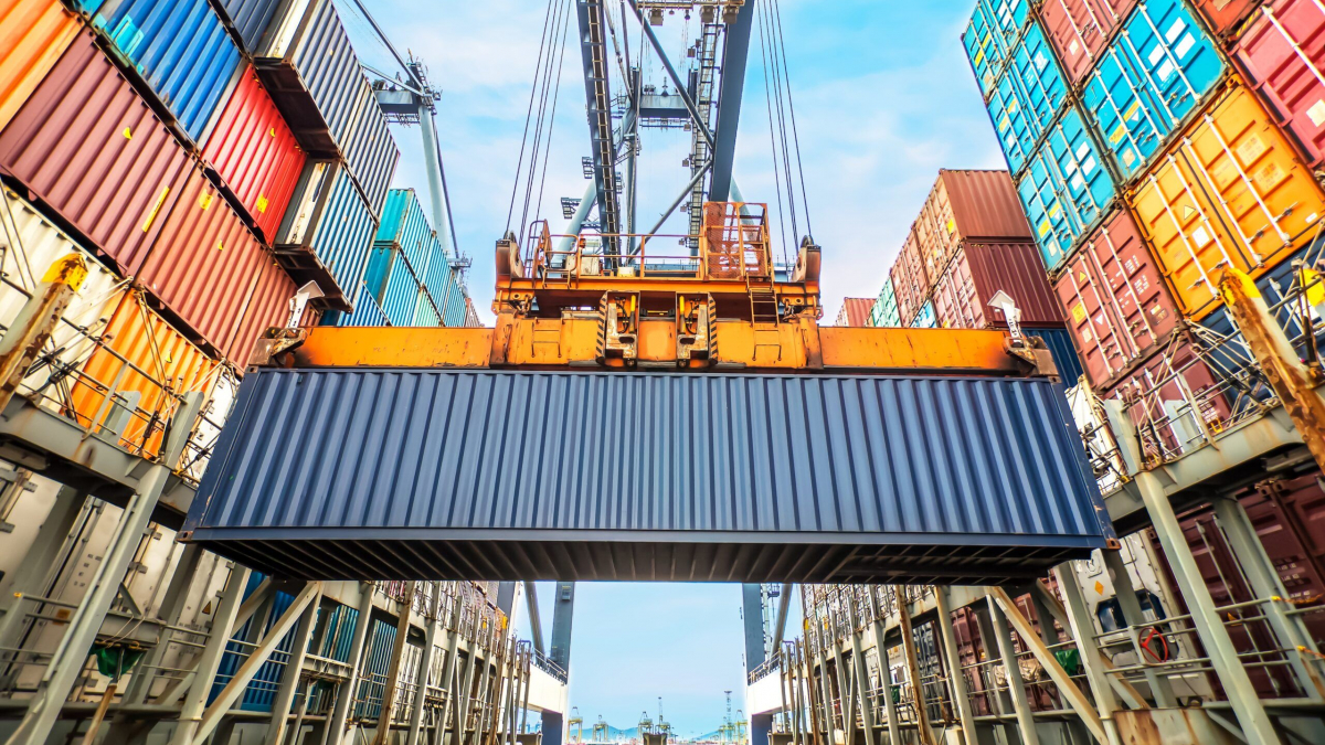 A large crane lifts a blue shipping container above a cargo ship, surrounded by stacks of colorful containers under a bright sky at a busy port.