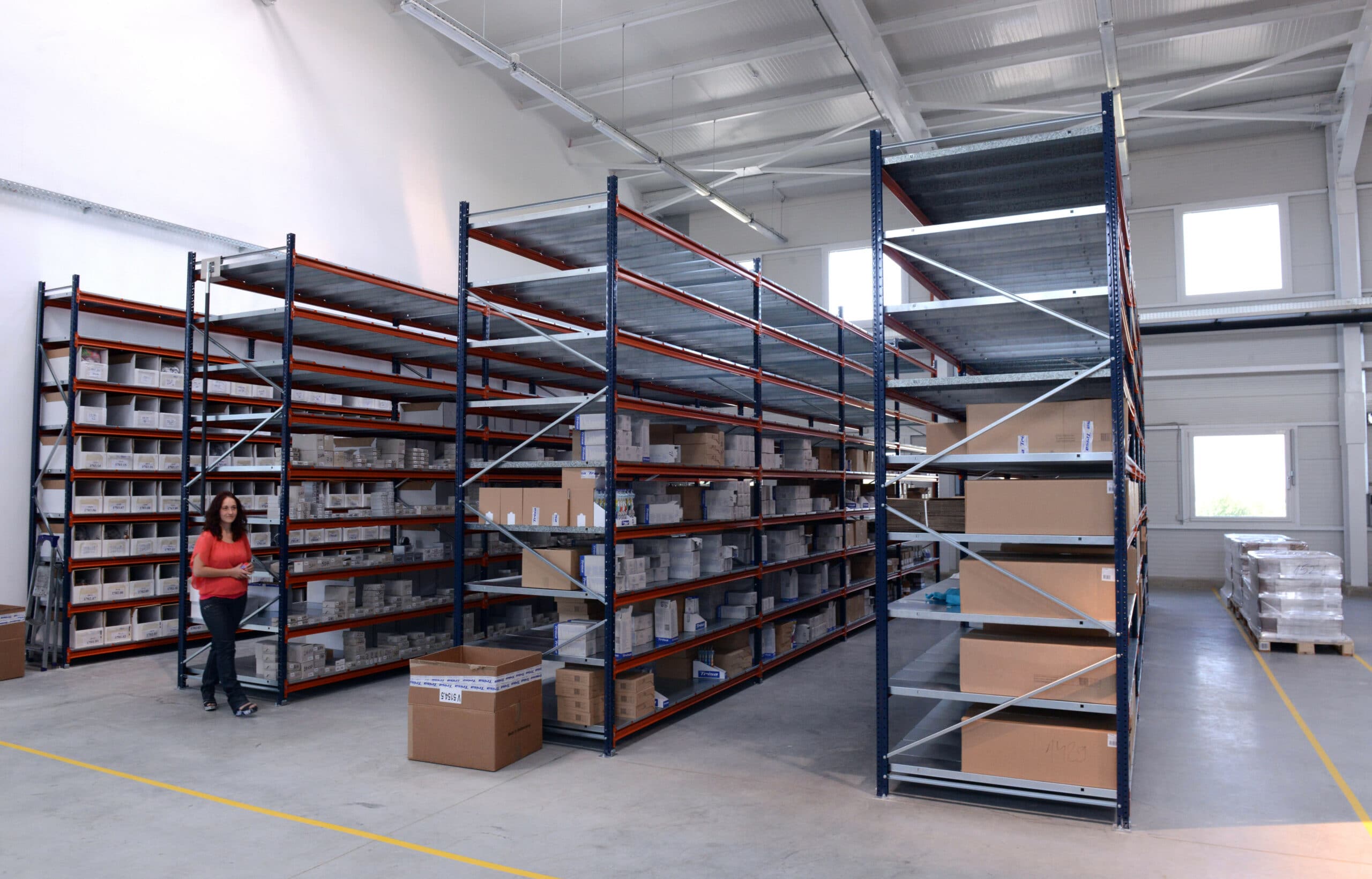 A woman walks through a spacious warehouse with tall metal shelves filled with boxes and packages. The floor is clean and there are additional boxes on the ground near the shelves.